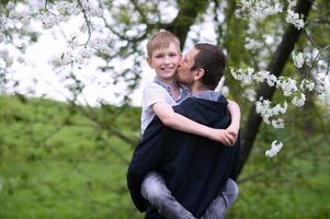 le garçon dans papa bras regards dans le caméra et sourires, papa baisers le sien fils photo