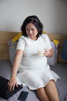 content Jeune asiatique Enceinte femme en buvant Lait sur lit à Accueil tandis que en portant sa ventre et en pensant photo
