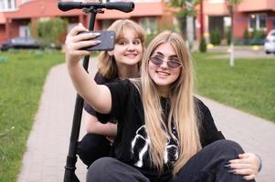 deux marrant les filles sont séance sur une scooter dans le ville et prise selfies sur leur téléphone. adolescents sourire photo