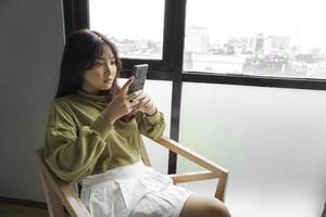 une Jeune asiatique femme est sentiment sérieux et concentré sur en portant sa téléphone intelligent dans main tandis que séance dans une chambre à coucher. photo