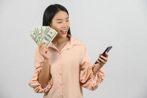 portrait de une de bonne humeur Jeune femme en portant argent billets de banque et en utilisant téléphone intelligent isolé plus de blanc Contexte photo