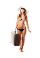 plein longueur portrait de une magnifique Jeune femme posant dans une bikini, chapeau et des lunettes de soleil avec une valise dans main sur blanc photo