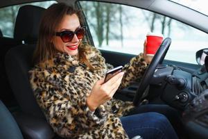 femme d'affaires dans une fourrure manteau avec rouge lèvres Envoi en cours une texte message et en buvant café tandis que conduite photo