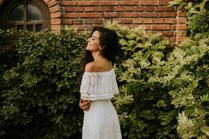 une Jeune femme avec frisé cheveux des stands dans une magnifique jardin, habillé dans une blanc écoulement robe photo