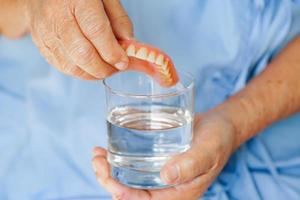 asiatique Sénior femme patient nettoyer les dents dentier dans une verre avec Solution pour mâcher aliments. photo