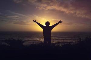 silhouette de une homme en train de regarder Orange le coucher du soleil sur falaise plus de le mer avec ouvert bras photo
