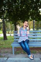 une portrait de une souriant femme dans une parc sur une banc parlant sur le téléphone photo