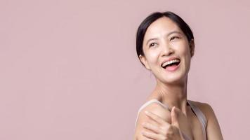 magnifique Jeune asiatique femme content sourire avec nettoyer visage peau dans marron soutien-gorge, spa costume sur rose Contexte. soins de la peau, traitement, bien-être thérapie, faciale se soucier, beauté femelle santé, cosmétologie concept photo