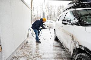 homme lavant une voiture suv américaine à eau haute pression avec galerie de toit au lavage en libre-service par temps froid. photo