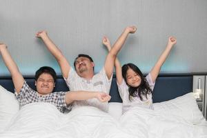 content famille réveiller en haut dans Matin élongation main monter en haut à le air tandis que séance sur lit dans chambre photo
