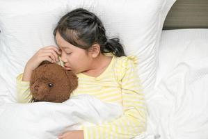 sucré peu fille est en train de dormir avec une nounours ours dans sa lit à maison, sucré rêver photo