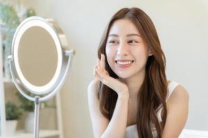 frais, agréable en bonne santé peau, magnifique de asiatique Jeune femme, fille à la recherche à miroir, émouvant sa visage avec avant faire en haut cosmétique routine à maison. femelle Regardez avec Naturel mode style, faciale beauté. photo