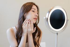 frais, agréable en bonne santé peau, magnifique de asiatique Jeune femme, fille à la recherche à miroir, émouvant sa visage avec avant faire en haut cosmétique routine à maison. femelle Regardez avec Naturel mode style, faciale beauté. photo