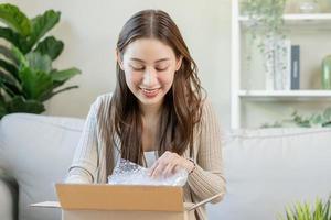 content excité, asiatique Jeune femme, fille client séance sur canapé à maison, ouverture et déballage papier carton boîte carton parcelle après achat commande cadeau, achats en ligne, livraison un service concept. photo