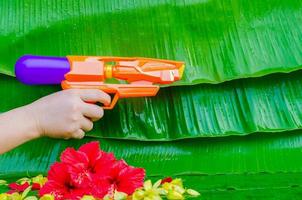 main en portant l'eau pistolet avec coloré fleurs sur humide banane feuilles Contexte pour Thaïlande Songkran festival. photo