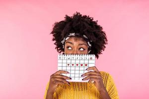 portrait de une marrant Jeune africain américain fille dans cache derrière une menstruel périodes calendrier et à la recherche une façon à copie espace isolé plus de rose Contexte. femelle période calendrier photo