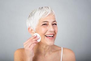 en bonne santé mature femme enlever maquillage de sa visage avec coton tampon isolé sur gris Contexte. beauté portrait de content femme nettoyage peau et à la recherche à caméra. photo