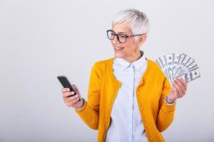 portrait de une Enchanté Sénior femme en portant bouquet de argent billets de banque et payant en ligne sur sa mobile téléphone plus de gris Contexte. content Sénior femme en portant beaucoup de espèces. photo
