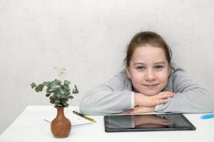 peu mignonne fille séance à le table dans de face de le tablette avec sa mains plié et souriant, apprentissage photo