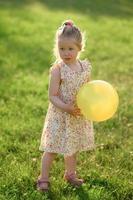 peu fille 3 ans vieux dans une été robe dans une clairière avec un Jaune Balle dans sa mains. le fille est ne pas content photo