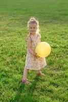 peu fille 3 ans vieux dans une été robe dans une clairière avec un Jaune Balle dans sa mains. le fille est heureux. photo