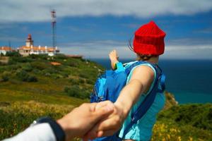 suivre moi - content Jeune femme dans une rouge chapeau et avec une sac à dos derrière sa retour tirant gars main. main dans main en marchant à le phare photo