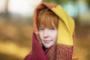 une roux fille enveloppé se dans une de laine couverture dans un l'automne parc. enfant dans l'automne. photo