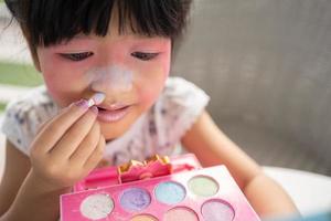 adorable peu enfant asiatique fille des peintures sa bouche avec rose les enfants têtes et regards dans le miroir. une enfant pièces à Accueil dans une jouet beauté salon. augmenter apprentissage développement pour enfants d'âge préscolaire. photo