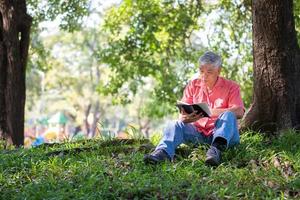 content asiatique vieux Sénior homme avec gris cheveux en train de lire livre à l'extérieur dans parc. concept de content personnes âgées homme après retraite et bien santé photo