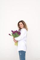mignonne fille dans une blanc chemise étreindre une grand bouquet de violet tulipes sur une blanc Contexte photo
