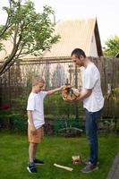 une mignonne garçon dans une blanc T-shirt recueille bois de chauffage et donne il à le sien père. bois de chauffage sur le herbe photo