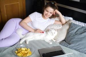 une mignonne fille est mensonge sur une lit avec une chat et à la recherche à une portable photo