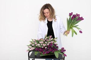 une mignonne fille prend une violet tulipe de le table pour une bouquet. des stands sur une blanc Contexte photo