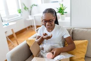 Jeune satisfait content africain fille femme Dame Shopaholic client asseoir sur canapé déballer parcelle livraison boîte, en ligne achats expédition concept. prise Photos de produit à Publier sur social médias