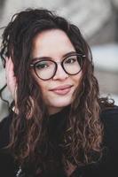 magnifique Jeune femme avec brunette frisé cheveux, portrait dans œil des lunettes profiter le Soleil dans le ville. photo