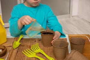 peu fille est arrosage le nouvellement planté graines, sur le table sont une râteau, une pelle, tourbe marmites, plantation semis photo