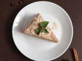 Haut vue de délicieux napoléon gâteau avec crème sur une blanc assiette décoré avec une brin de menthe sur une marron Contexte. une cannelle bâton, Badyan, café des haricots sur une marron Contexte. copie espace photo