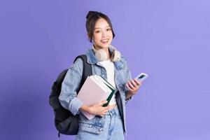 portrait de une magnifique asiatique écolière portant une sac à dos sur une violet Contexte photo