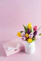 magnifique bouquet de coloré tulipes dans une blanc céramique vase sur une rose Contexte avec une de fête cadeau boîte. verticale voir. une copie espace. photo