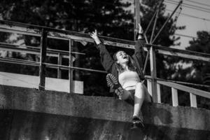 une touristique femme dans décontractée vêtements et été des sandales est assis sur Haut de le chemin de fer dans le soir à coucher de soleil, élevé sa mains et se réjouit, noir et blanc photo