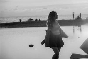 une magnifique touristique femme avec longue ondulé cheveux dépense été temps dans le soir à le coucher du soleil sur le rivage, le fille danses sur le océan. noir et blanc photo. photo