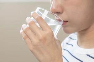 content beau, asiatique Jeune femme, fille en buvant, siroter Frais verre de l'eau pour hydratation de corps, en portant transparent verre dans sa main, soif à maison. santé se soucier, en bonne santé mode de vie concept. photo