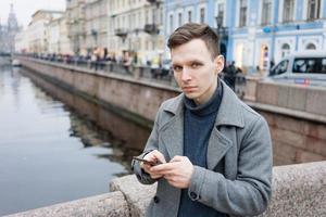 Beau Jeune homme en utilisant mobile téléphone, portant un élégant gris manteau, des stands photo