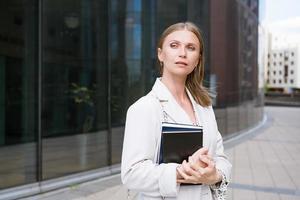 réussi affaires femme détient une carnet dans sa main dans de face de un Bureau bâtiment. jolie photo