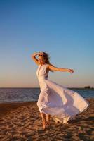 magnifique fille dans une longue blanc robe par le mer dans le soir à le coucher du soleil photo