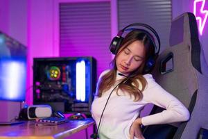 Jeune gens mal au dos. joueur et e-sport en ligne de asiatique femme en jouant en ligne ordinateur vidéo Jeu avec éclairage effet, diffuser diffusion vivre à maison. elle une mal au dos dû à séance pour une longue temps photo