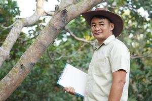 Beau asiatique homme botaniste tenir papier presse-papiers, des stands à côté de une arbre dans forêt. concept, enquête ,recherche botanique les plantes. forêt et environnement conservation. photo