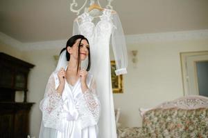 le Matin de le la mariée. portrait de une brunette la mariée dans une blanc soie manteau derrière une robe photo
