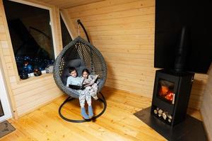 frère avec sœur en train de lire livres dans confortable pendaison chaise contre cheminée dans confortable en bois minuscule cabine maison. la vie dans campagne. photo