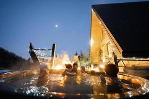 famille profiter baignade dans en bois baril chaud baignoire dans le terrasse de le chalet. scandinave baignoire avec une cheminée à brûler bois et chaleur l'eau. photo